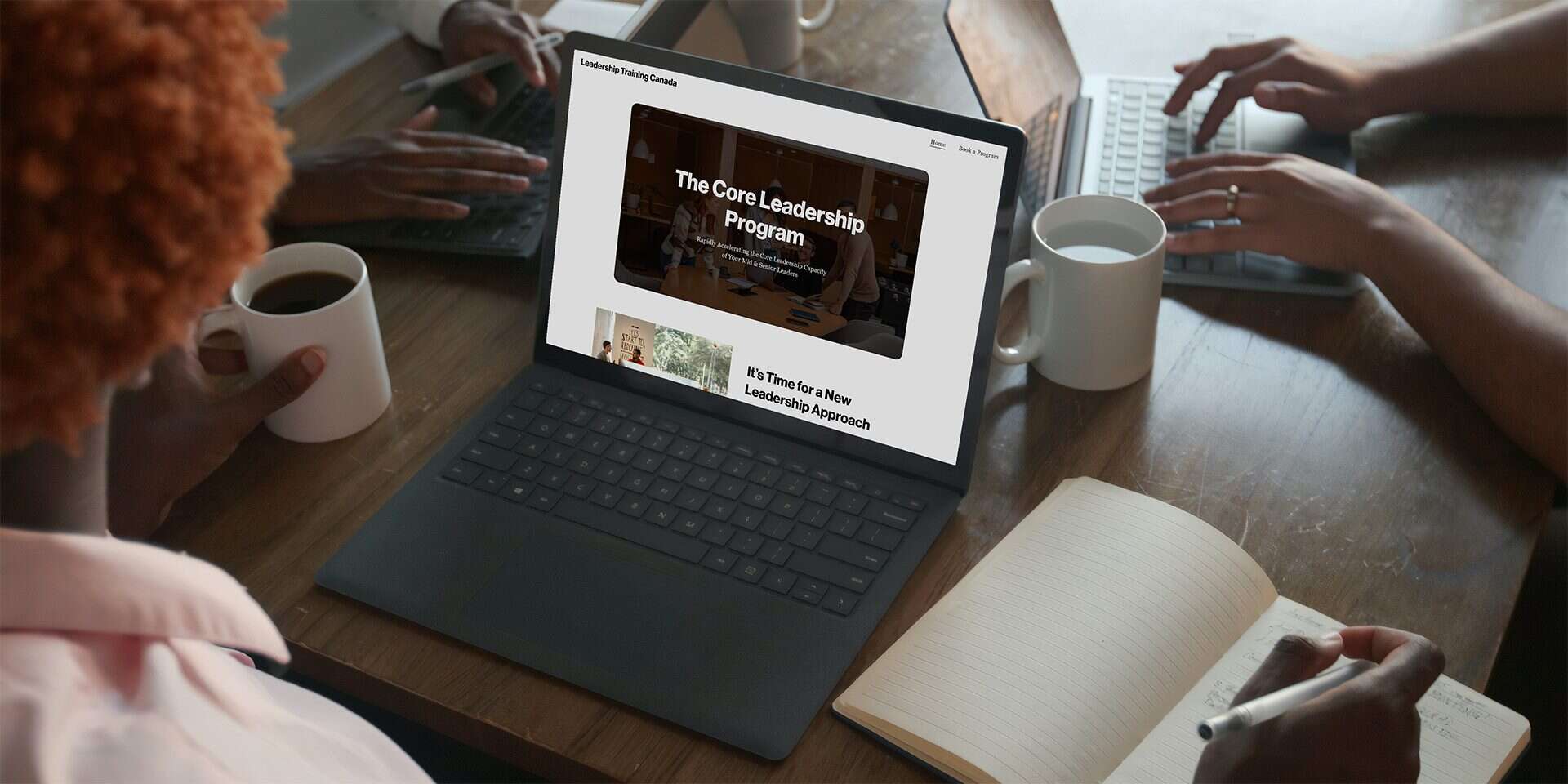 Leadership Training Canada homepage open on a laptop on a busy table with coworkers meeting around.